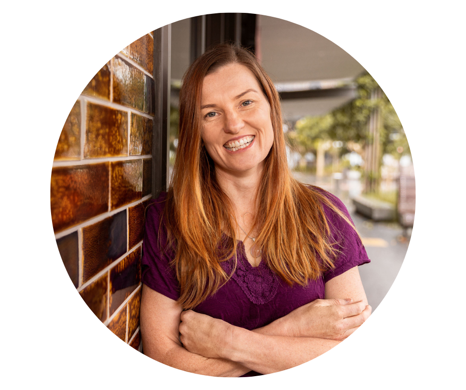 Anj Young, Clinical Director at Top Notch, smiling with her arms crossed, leaning against a tiled wall. She is wearing a purple shirt and standing outdoors with trees and buildings in the background